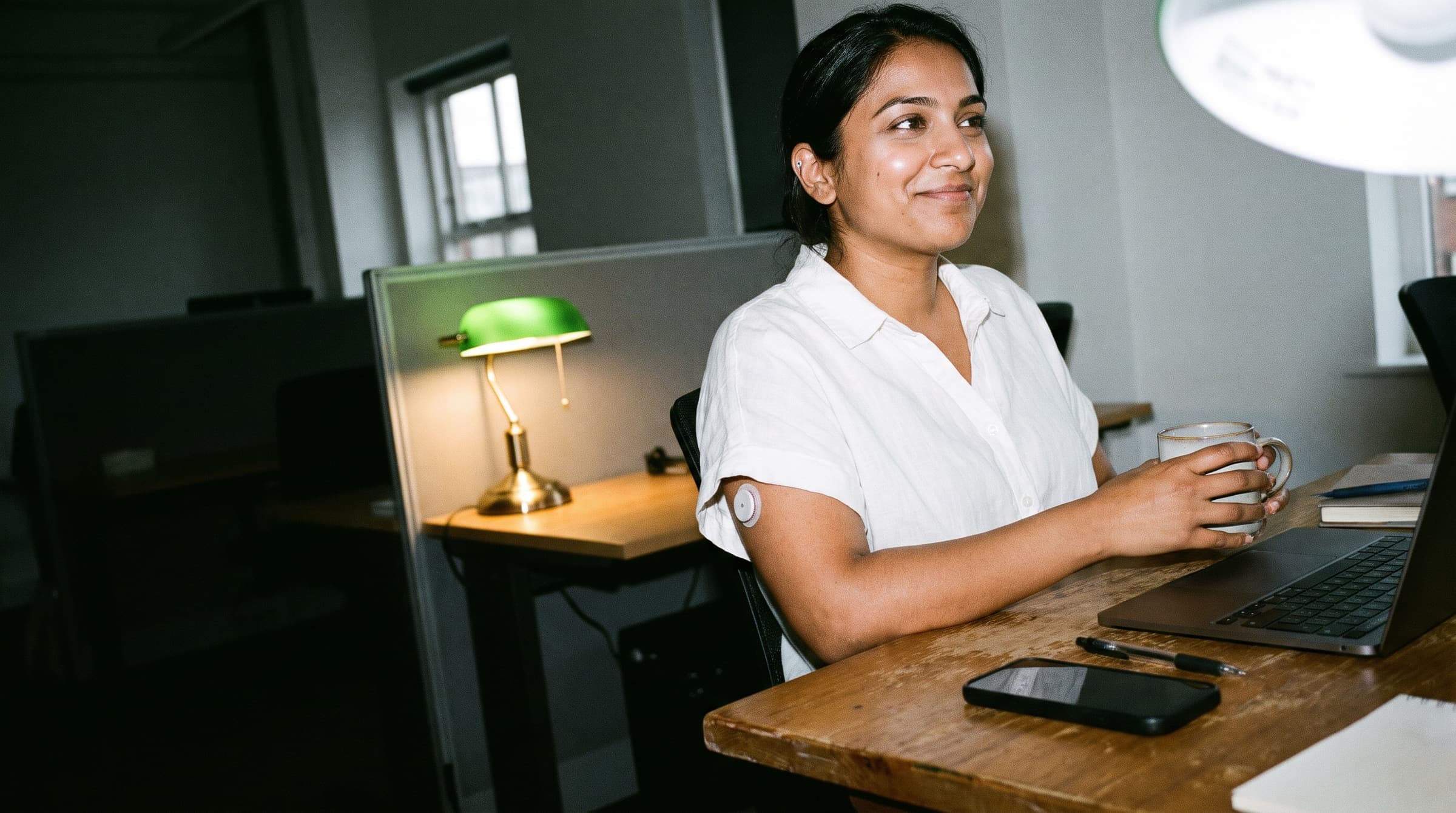 A woman at her office desk in the afternoon, smiling with a mug in hand, an energy sensor on her upper arm