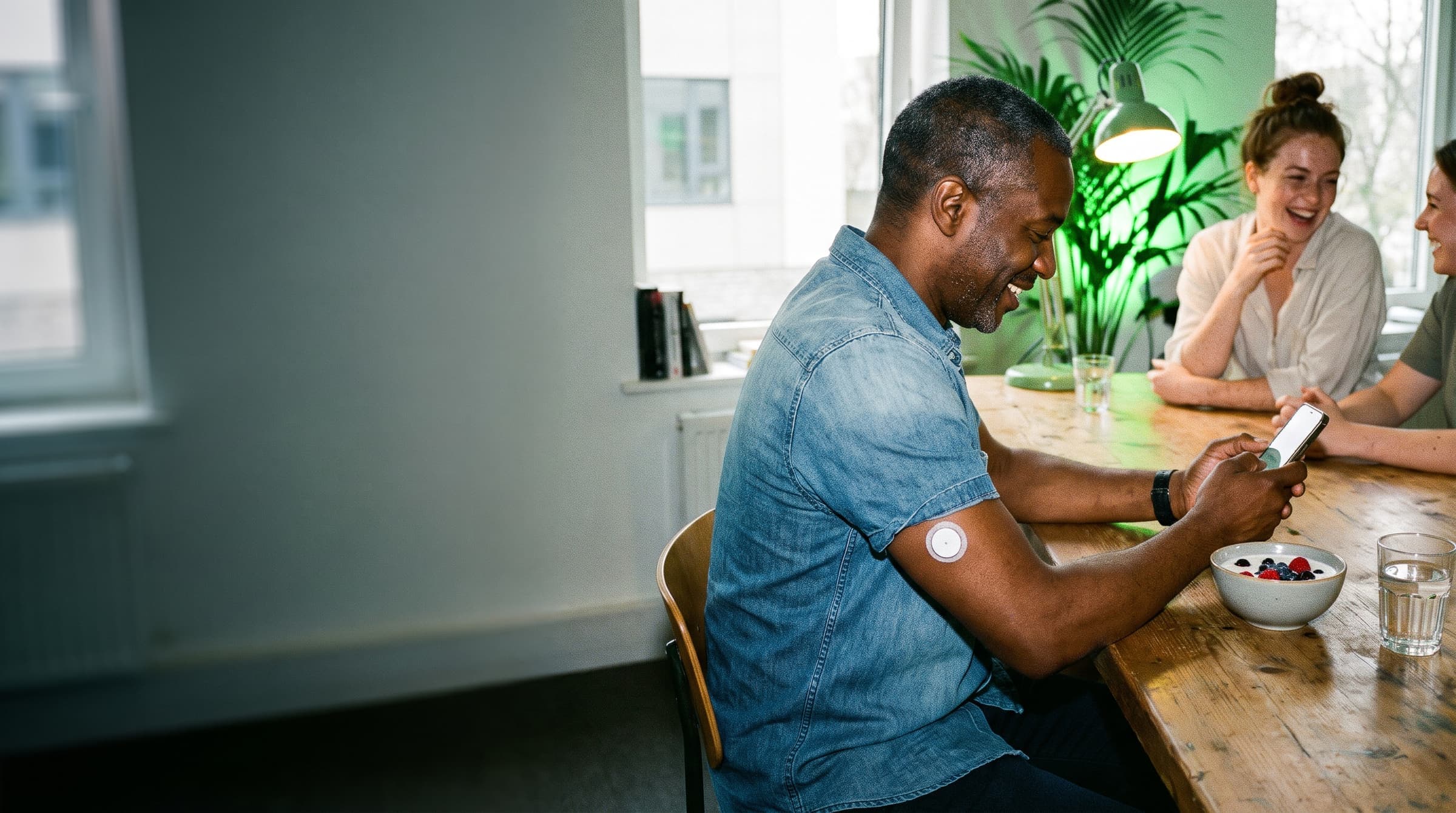 A man smiling at his phone over breakfast at a shared office kitchen table while colleagues laugh beside him, an energy sensor on his upper arm