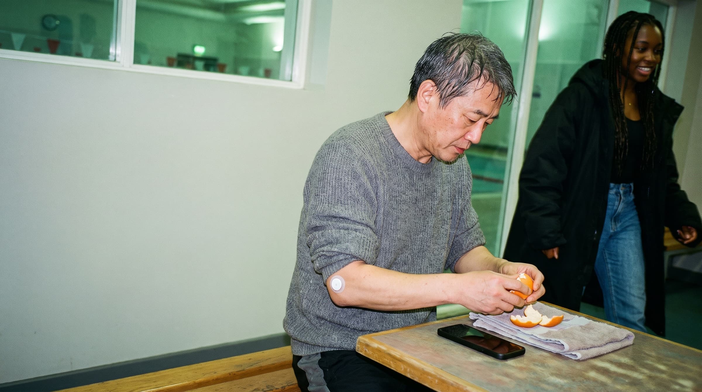 A man peeling an orange in a swimming club café after a morning swim, an energy sensor on his upper arm