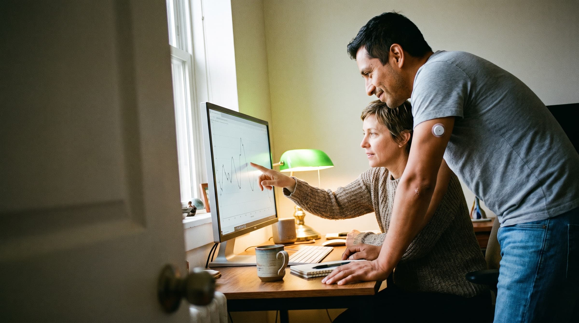 A practitioner pointing at a glucose line on a large desktop screen while her client leans in, an energy sensor on his upper arm
